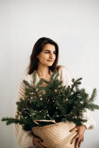 Smiling young woman in a cozy white sweater holding a decorated Christmas tree in a basket against a white wall.