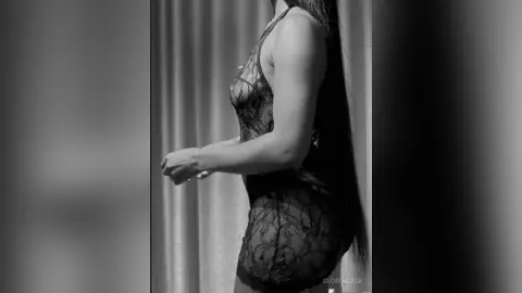 Side view of a woman in a black lace bodysuit standing in a dimly lit room.