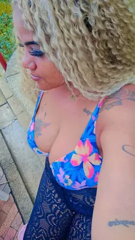 Blonde woman in a blue floral bikini top and black lace skirt posing outdoors in sunny weather.