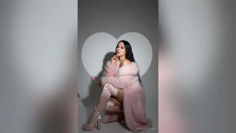 AnnyeWilliams poses in a fluffy pink fur coat and matching boots against a heart-shaped shadow background while holding a pink lollipop.