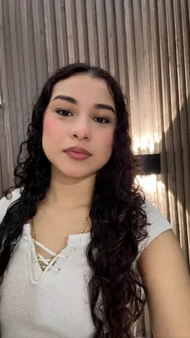 Young woman with long curly hair wearing a white waffle knit top in a warm, modern indoor setting.