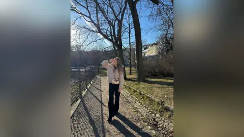MargaritaHafele stands on a path in a park, wearing a light tan jacket and black pants, with bare arms and blonde hair under a clear blue sky.