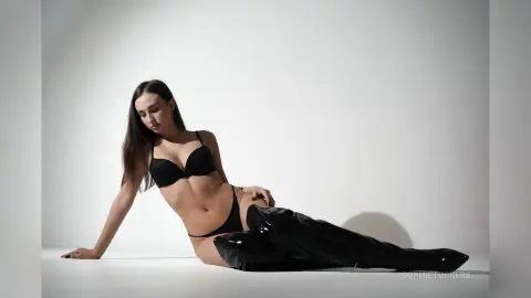 Young woman in black lingerie and high-gloss thigh-high boots posing against a minimalist white studio background.