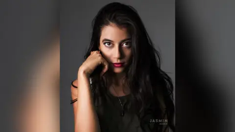 JessieLiu poses with her hand against her chin, wearing a dark olive tank top and red lipstick with long, wavy black hair.