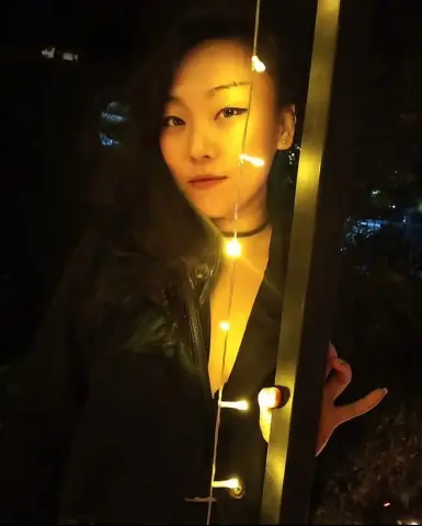 Young woman in a black outfit posing by glowing fairy lights at night for a moody aesthetic portrait.