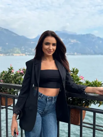 Stylish woman in a black blazer and denim jeans posing by a scenic lake under a blue sky.
