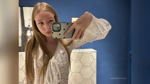 AdenaMcfalls wearing a white lace long-sleeve top, taking a mirror selfie with her blonde hair down against a blue wall.
