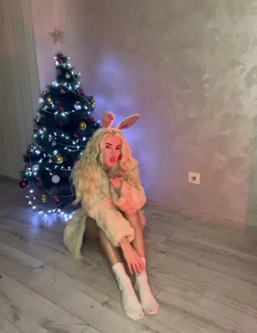 Blonde woman in a fuzzy bunny costume sitting by a glowing Christmas tree in a cozy festive room.