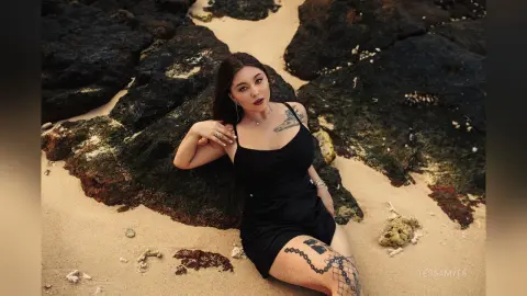 Young woman in a black tank top relaxing on a rocky beach under warm sunlight.