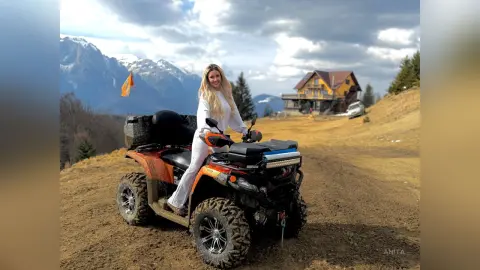 Anita sits upright on an orange ATV in a muddy field with mountains in the background, wearing a white long-sleeve top and white pants.