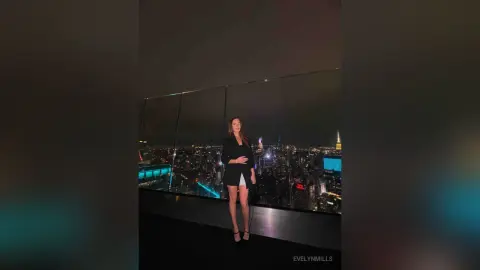 EvelynMills stands on a balcony overlooking a bright city skyline at night, wearing a black dress and black heels.