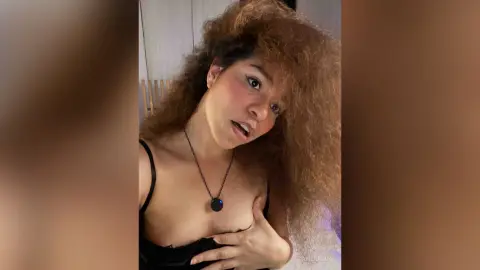 Young woman with voluminous curly hair wearing a black camisole and necklace in a dimly lit indoor setting.