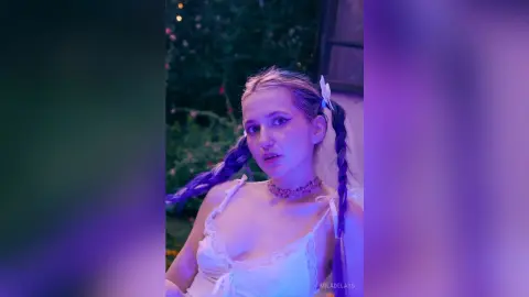 Young woman in a white lace dress with braided hair posing in a dreamy purple-lit garden setting.
