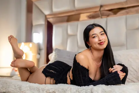 Smiling young woman in black loungewear relaxing on a cozy white bed in a bright, modern bedroom.