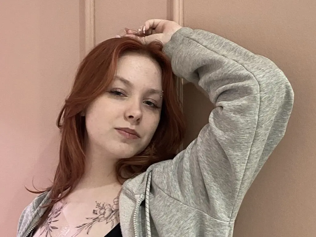 Redheaded woman in a grey oversized hoodie leaning against a pink wall with a relaxed, moody expression.