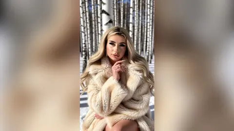 Blonde woman in a luxurious cream faux fur coat posing in a snowy birch forest with a glamorous mood.