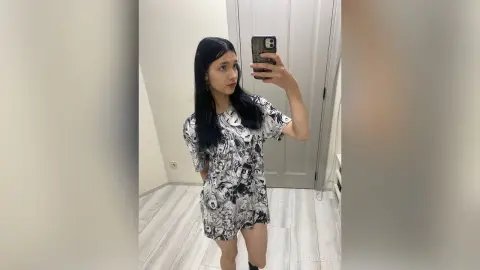 Young woman in a black and white graphic print mini dress taking a mirror selfie in a bright room.