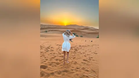 ChristineDavis stands in a desert landscape at sunset, wearing a short white dress and a headwrap while looking out over the sand dunes.