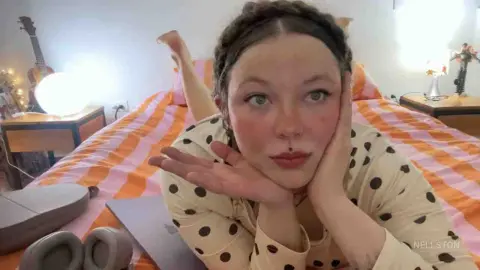 Young woman in a polka dot shirt resting her chin on her hands on a striped bed in a cozy bedroom setting.