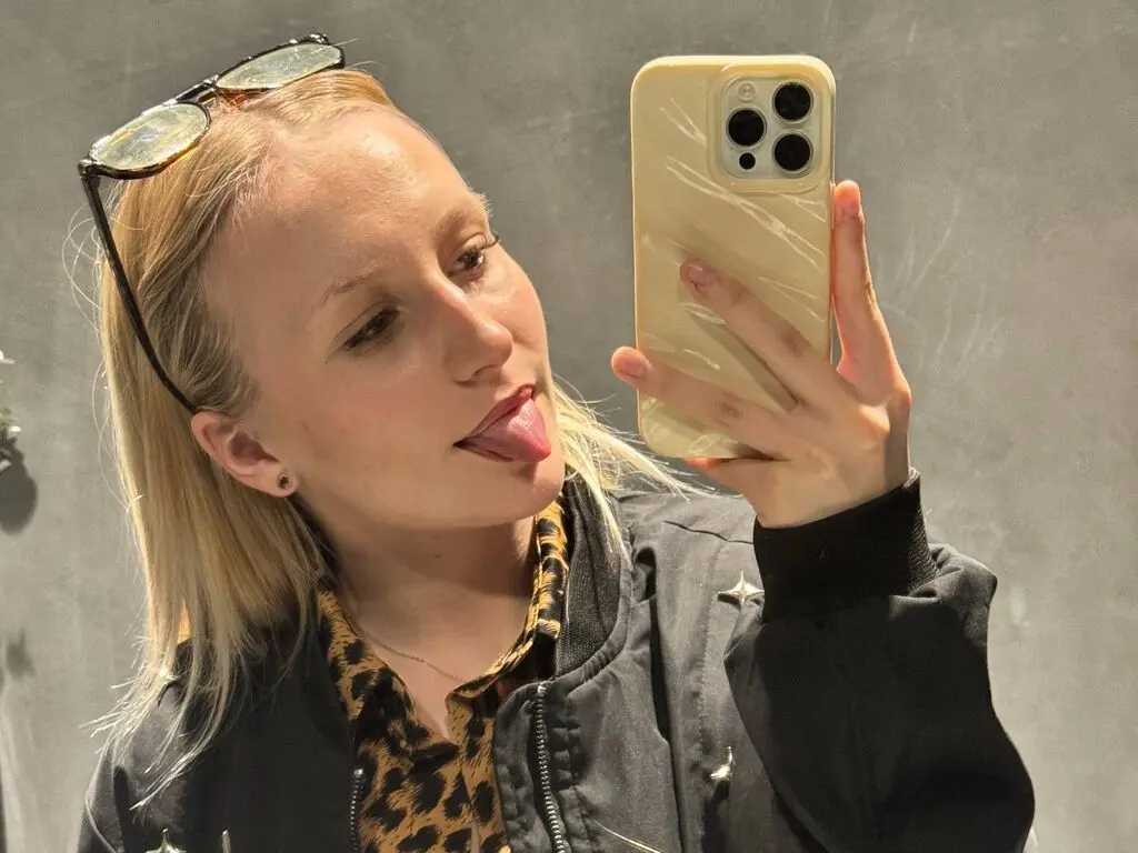Blonde woman in a leopard print shirt and black jacket taking a playful mirror selfie in a neutral setting.