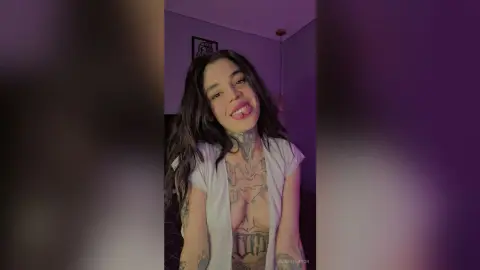 Smiling young woman with long dark hair and tattoos wearing a white sleeveless top in a dimly lit room.