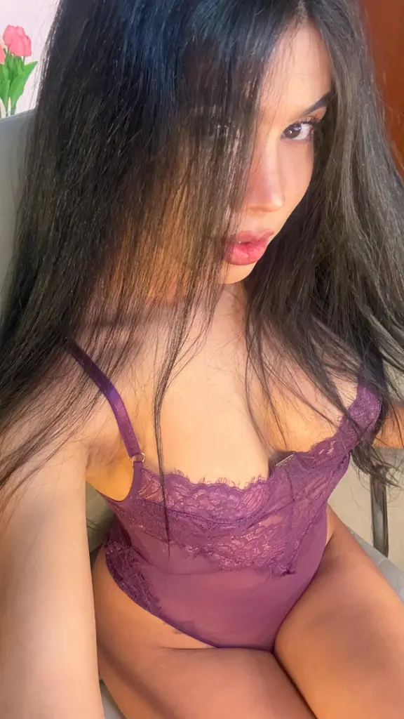 LittleMoon111 poses with long dark hair, wearing a black button-down shirt that shows cleavage, set against a dark curtain background.