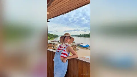 TefyRusso wearing a tan sun hat and red and white striped tank top with denim overalls, posing outdoors by a river and wooden balcony.