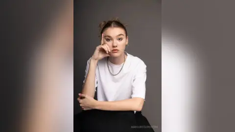Young woman in a white t-shirt and silver necklace posing thoughtfully against a gray studio background.