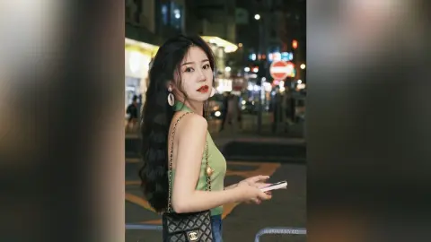 Young woman in a green tank top holding a phone on a city street at night with glowing urban lights.