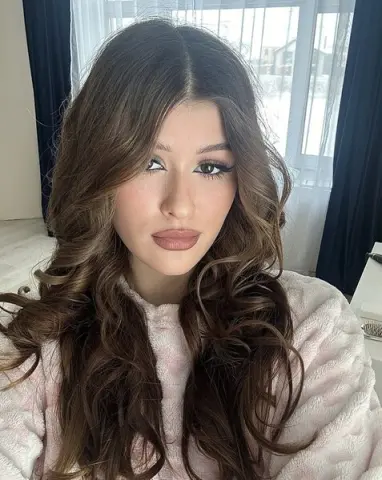 Young woman with long wavy brown hair and glam makeup wearing a cozy pink sweater in a bright room.
