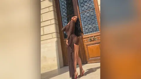 NicoleBella stands against a large wooden door wearing a tight brown long-sleeve dress with a high slit and red high heels.
