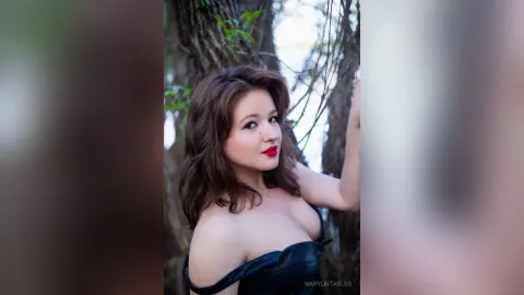 Beautiful woman in a black off-the-shoulder dress posing in a dreamy, sunlit forest setting.