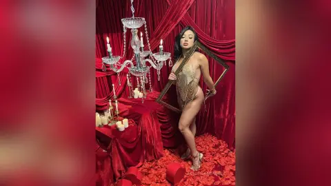 Glamorous woman in a gold sequin bodysuit posing by a crystal chandelier against red velvet curtains.