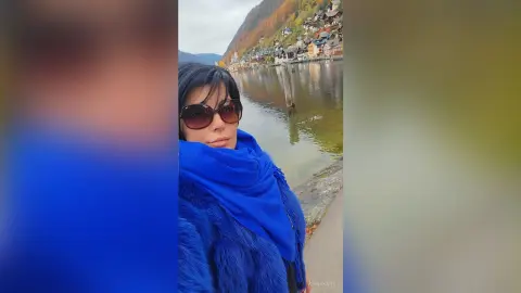 Woman in a blue winter coat and sunglasses smiling by a scenic lake and mountain village.