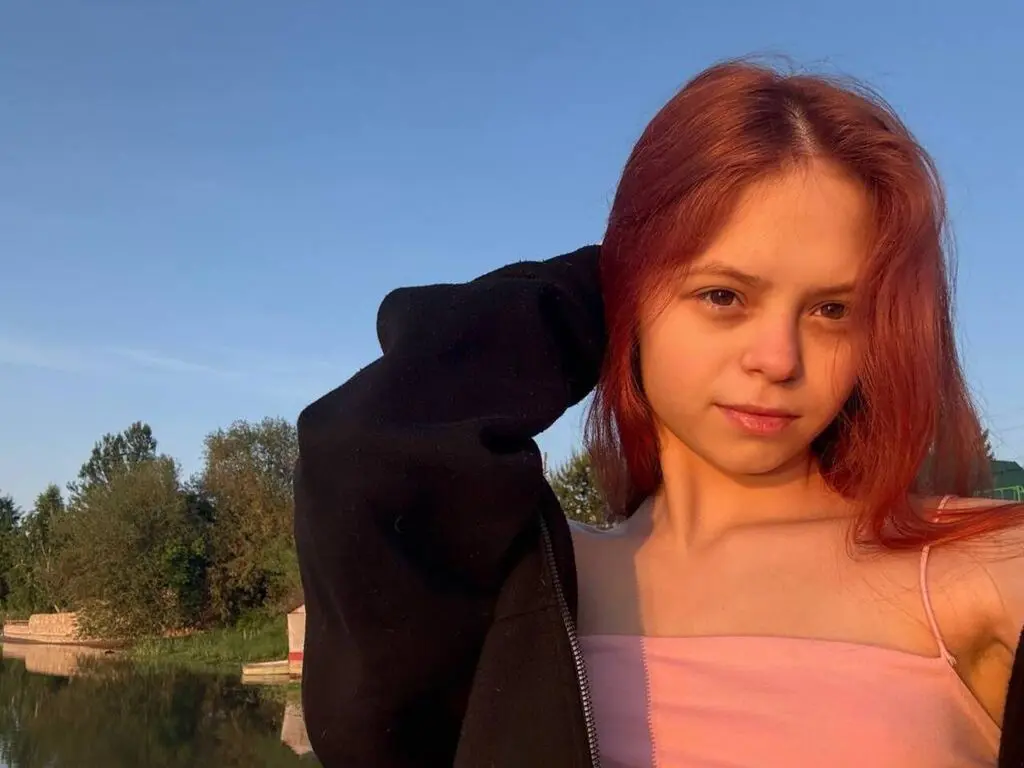 Young woman with red hair in a pink camisole posing outdoors against a sunset sky and trees.