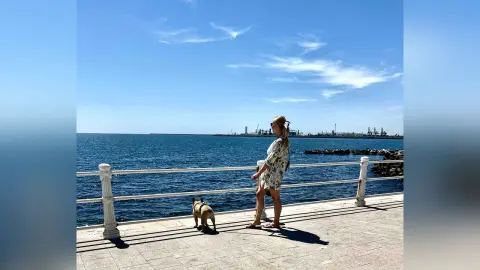 Woman in a floral summer dress walking her dog along a sunny seaside boardwalk under a blue sky.