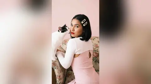 Stylish woman in a pink ribbed dress cuddling a black and white cat in a cozy, pastel-toned room.