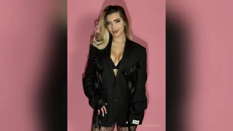 Elegant woman in a black blazer and pearl necklace posing against a pink background with a sophisticated mood.