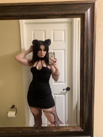 BimboKitten poses in a black mini dress and fishnet stockings, wearing black cat ears and holding a phone for a mirror selfie.