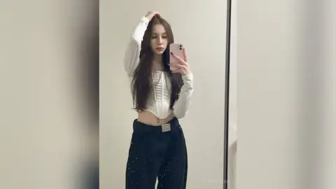Young woman in a white long-sleeve crop top and dark trousers taking a mirror selfie in a bright room.