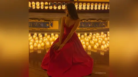 Elegant woman in a flowing red gown sitting amidst glowing candles in a warm, romantic ballroom setting.
