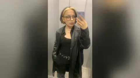 Trendy woman with blonde highlights wearing a black leather jacket and glasses taking a mirror selfie.