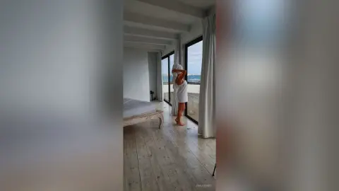 AnnaBanks stands in a bright room wearing a white towel, posing near a glass door with a view of the ocean.