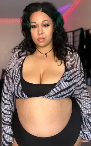 Curvy woman in black bikini and patterned mesh cardigan posing in a room with pink neon lighting.