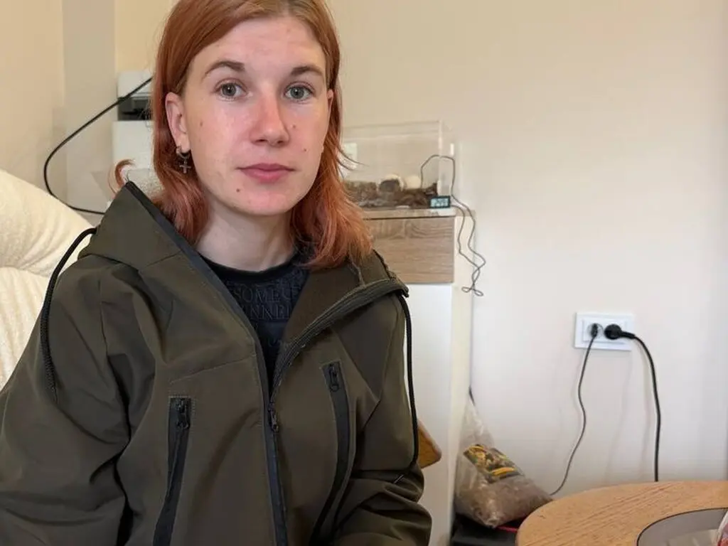 Red-haired woman in an olive green jacket sitting in a bright indoor room with a calm expression.
