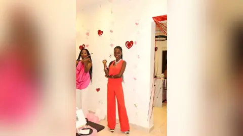 Smiling Black woman in a vibrant red jumpsuit posing in a room decorated with red heart balloons.