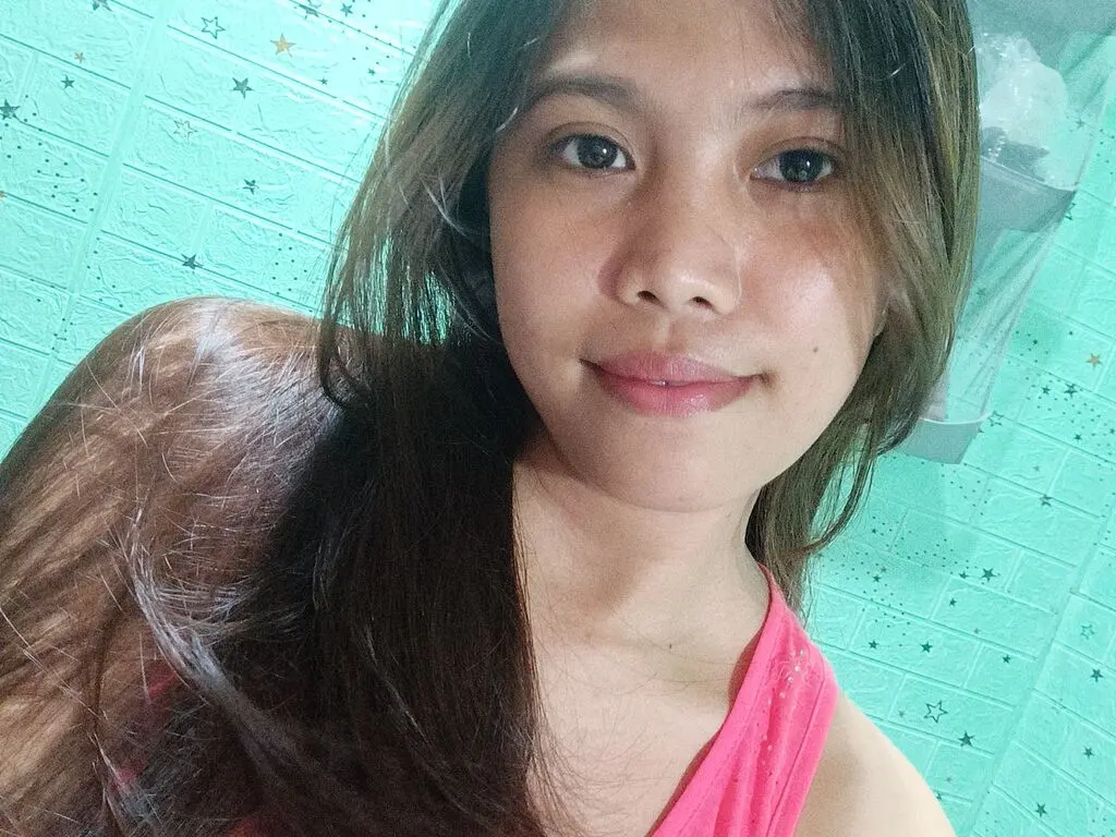 Smiling young woman with long brown hair wearing a pink tank top in a bright teal room.