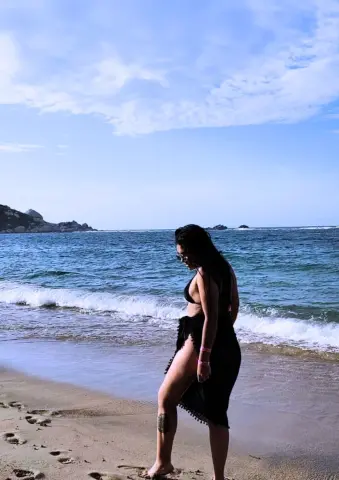 Woman in a black bikini and sarong walking on a sunny beach by the blue ocean waves.