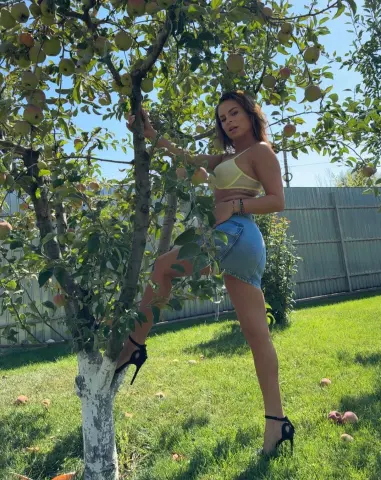 Young woman in denim shorts and crop top posing in a sunny apple orchard.