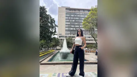 Stylish woman in a white crop top and black wide-leg trousers standing by a city fountain near a tall building.